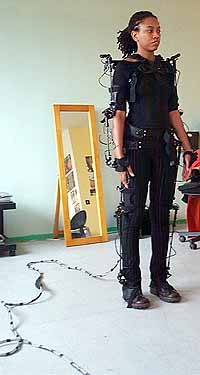 A woman wearing the exoskeletal Gypsy motion-capture suit, black straps and sensors down her limbs, cables trailing across the floor toward a mirror