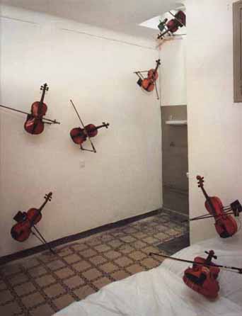 Installation photograph: red violins with their bows mounted swarming up white walls and across a tiled floor, like an infestation of insects