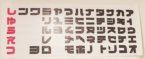 Wall chart of the katakana syllabary in a blocky graphic typeface, black characters in columns with red script down the left edge