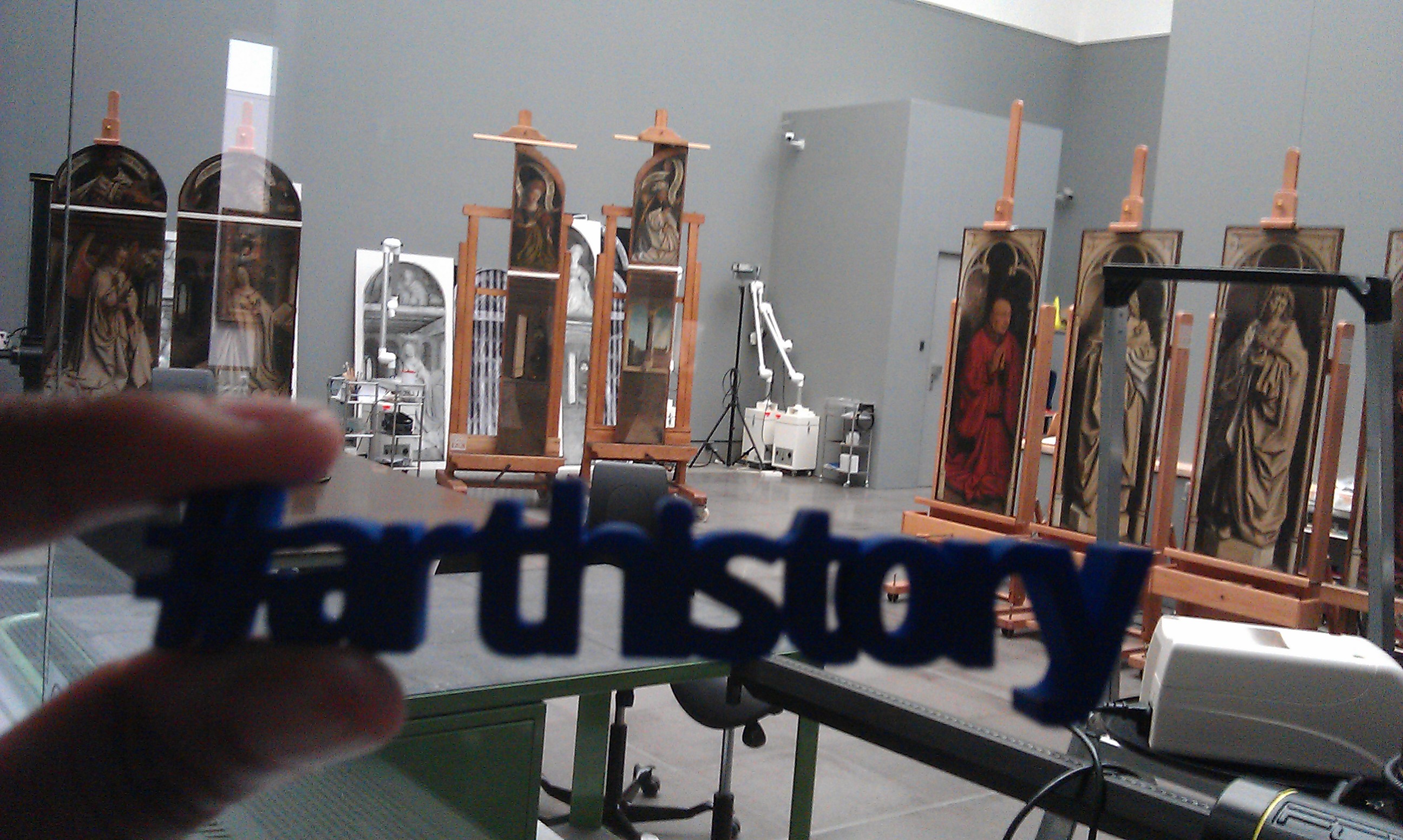 In the Ghent Altarpiece restoration studio: Van Eyck panels on easels, a hand holding up a blue 3d-printed hashtag arthistory in the foreground In the Ghent Altarpiece restoration studio: Van Eyck panels on easels, a hand holding up a blue 3d-printed hashtag arthistory in the foreground