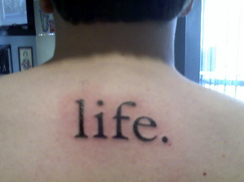 Photograph of the back of a neck and shoulders tattooed with the single lowercase word life, ending in a period
