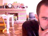 Webcam frame: a man's face looms half out of frame at the right; behind him a brick fireplace, a television, and a small posable skeleton bent double